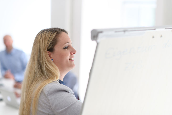 Frau steht neben einer FlipChart und erklärt etwas während Zuhörende aufmerksam folgen.