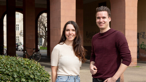 Zwei lächelnde Personen stehen nebeneinander vor einer Unterführung. Die Personen heißen Melina und Sandro.