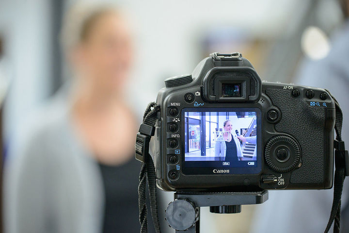 Auf der Bildanzeige einer Digitalkamera ist das Bild einer Frau zu sehen, die mit einem Mikrofon interviewt wird.