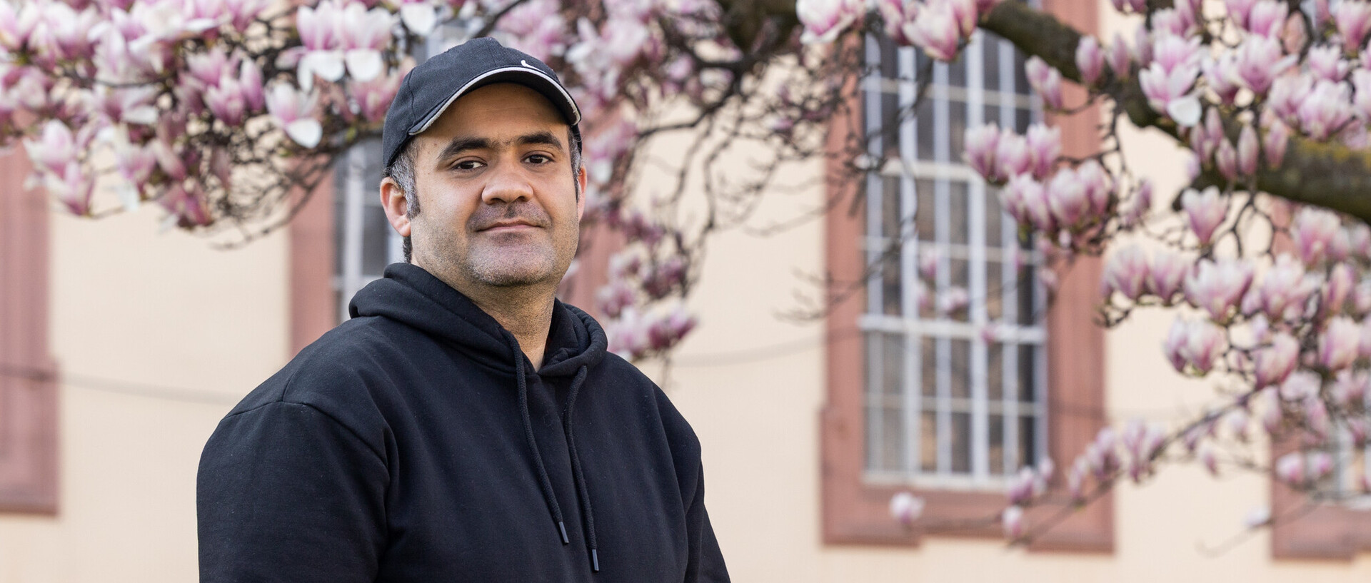 Porträt von Zubair, der vor einem Teil des Schlosses steht und in die Kamera schaut. Er trägt eine schwarze Kappe und einen schwarzen Hoodie. Im Hintergrund ist ein blühender Magnolienzweig zu sehen.