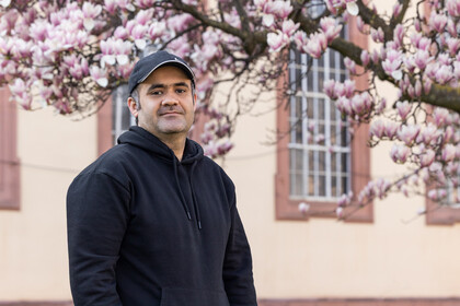 Porträt von Zubair, der vor einem Teil des Schlosses steht und in die Kamera schaut. Er trägt eine schwarze Kappe und einen schwarzen Hoodie. Im Hintergrund ist ein blühender Magnolienzweig zu sehen.