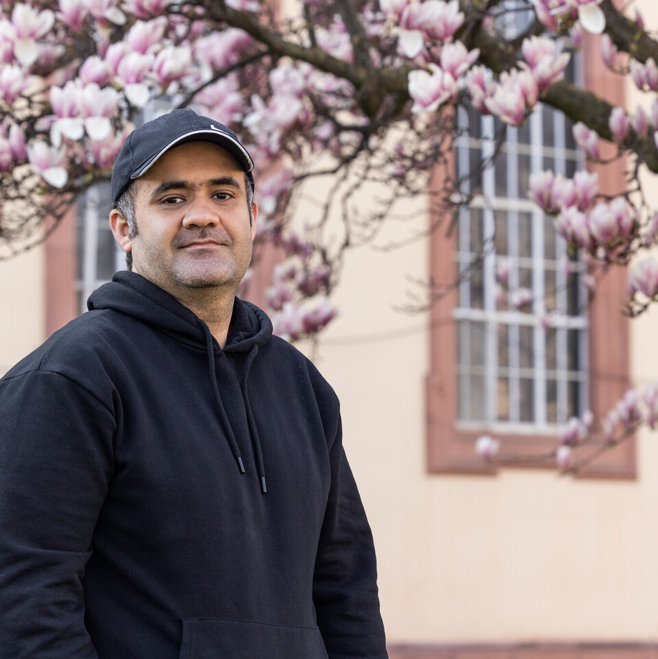 Porträt von Zubair, der vor einem Teil des Schlosses steht und in die Kamera schaut. Er trägt eine schwarze Kappe und einen schwarzen Hoodie. Im Hintergrund ist ein blühender Magnolienzweig zu sehen.