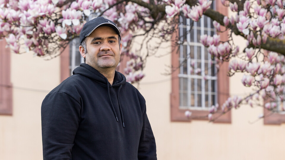 Porträt von Zubair, der vor einem Teil des Schlosses steht und in die Kamera schaut. Er trägt eine schwarze Kappe und einen schwarzen Hoodie. Im Hintergrund ist ein blühender Magnolienzweig zu sehen.