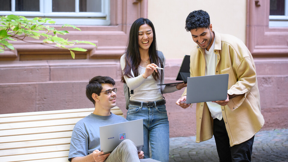 Drei Personen mit Laptops unterhalten sich lachend im Rektoratshof der Universität.