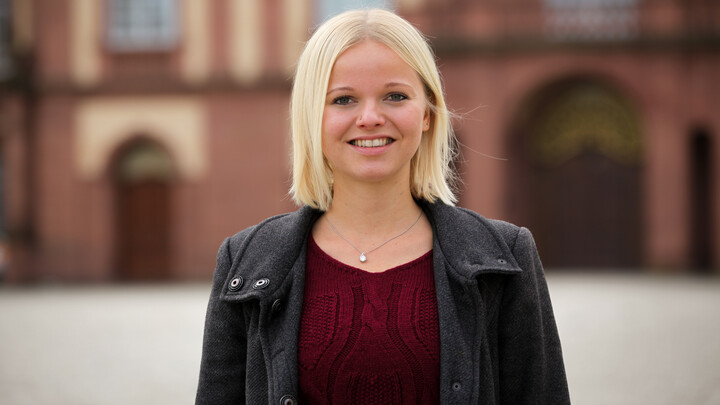 Katharina Siebenhaar. She is wearing a black leather jacket over a red shirt and is standing on the Ehrenhof in front of the castle.