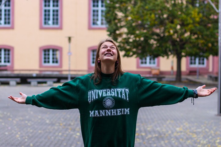 Eine Studentin in einem dunkel grünen Universität Mannheim Pullover steht mit ausgestreckten Armen im Schneckenhof und blickt nach oben.