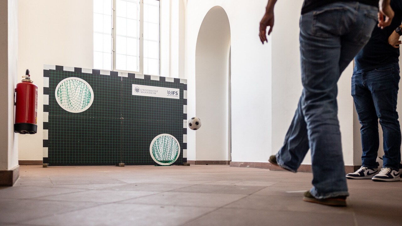 Eine Torwand mit zwei Löchern steht in den Katakomben. Rechts daneben in der Luft ein Fußball. Im Vordergrund auf der rechten Seite zwei Personen in Jeans. Die eine Person, von der die untere Körperhälfte sichtbar ist, trägt Jeans, steht mit dem Rücken zur Kamera und hat das rechte Bein angehoben. Die zweite Person, optisch verdeckt von der ersten, trägt ebenfalls Jeans.