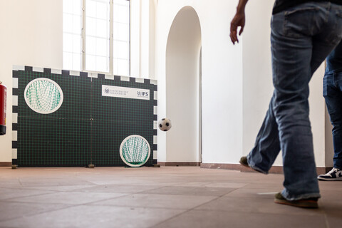 Eine Torwand mit zwei Löchern steht in den Katakomben. Rechts daneben in der Luft ein Fußball. Im Vordergrund auf der rechten Seite zwei Personen in Jeans. Die eine Person, von der die untere Körperhälfte sichtbar ist, trägt Jeans, steht mit dem Rücken zur Kamera und hat das rechte Bein angehoben. Die zweite Person, optisch verdeckt von der ersten, trägt ebenfalls Jeans. 