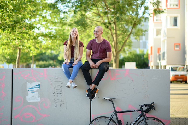Zwei Studierende sitzen lachend auf einer Betonmauer. An der Mauer lehnt ein Rennrad.