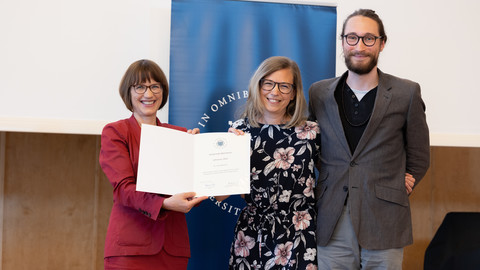 Drei Personen in schicker Klediung stehen nebeneinander auf einer Bühne. Die Person links hält eine Urkunde über einen Lehrpreis der Uni Mannheim an Lena Nadarevic in der Hand.