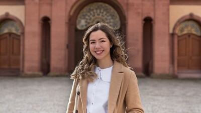 Eine lächelnde Person trägt eine weiße Bluse sowie einen hellbraunen Mantel und steht auf dem Ehrenhof vor dem Schloss. Die Person heißt Tsvetina Tsonkova. 