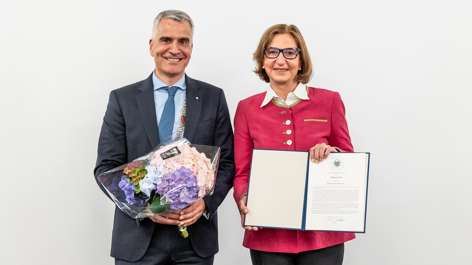 Margret Suckale, die einen roten Blazer, eine Brille und einen braunen Bob trägt, erhält die Ehrensenatorinnenwürde von Prof. Dr. Thomas Fetzer, der einen dunkelblauen Anzug mit blauem Hemd trägt. Beide stehen nebeneinander und lächeln in die Kamera. Prof. Dr. Fetzer hält einen Blumenstrauß.