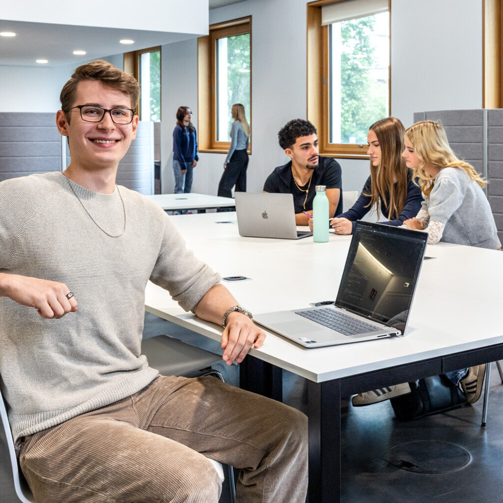 Ein Student sitzt mit einem Laptop an einem Gruppenarbeitstisch in B6. Im Hintergrund arbeiten 3 Studierende.