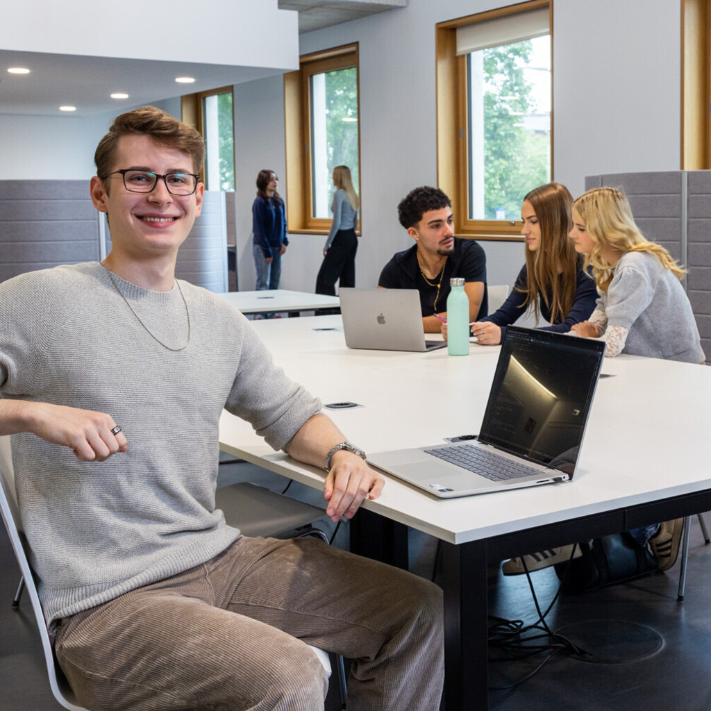 Ein Student sitzt mit einem Laptop an einem Gruppenarbeitstisch in B6. Im Hintergrund arbeiten 3 Studierende.