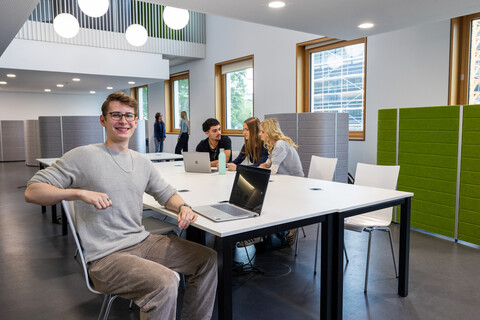 Ein Student sitzt mit einem Laptop an einem Gruppenarbeitstisch in B6. Im Hintergrund arbeiten 3 Studierende.