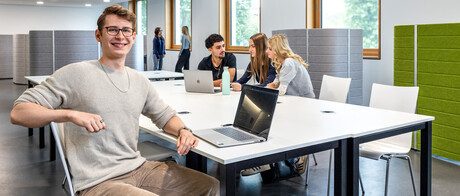 Ein Student sitzt mit einem Laptop an einem Gruppenarbeitstisch in B6. Im Hintergrund arbeiten 3 Studierende.