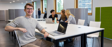 Ein Student sitzt mit einem Laptop an einem Gruppenarbeitstisch in B6. Im Hintergrund arbeiten 3 Studierende.
