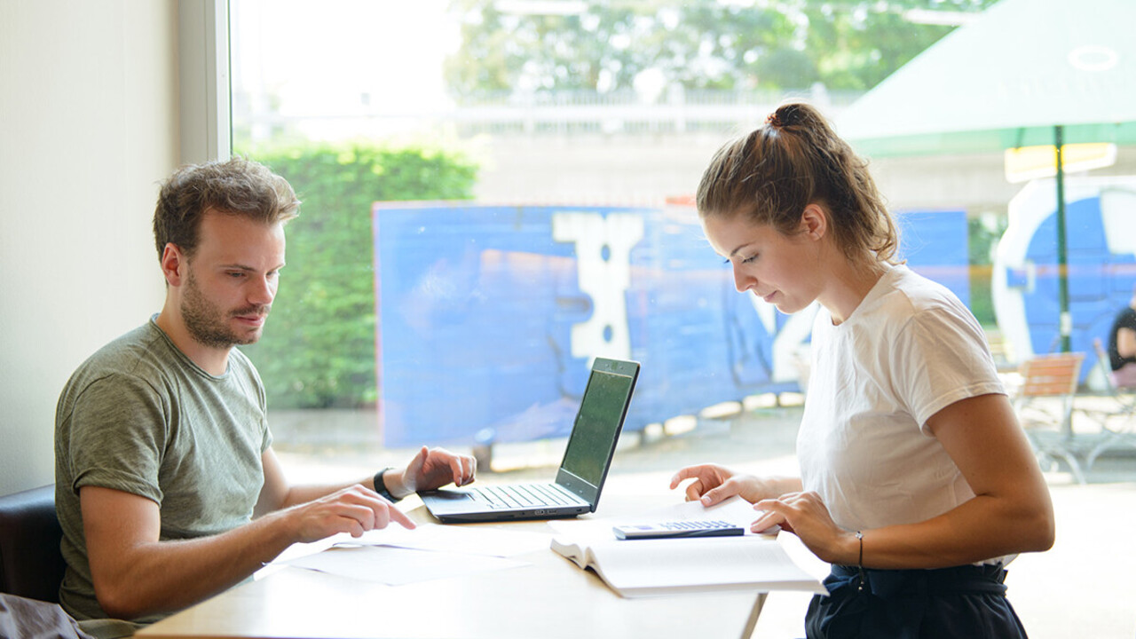 Eine Studentin und ein Student arbeiten gemeinsam an einem Tisch. Vor ihnen liegen ein Laptop, ein Taschenrechner und ein Buch.
