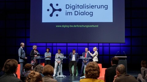 Sieben Personen sitzen und stehen nebeneinander um Stehtische verteilt und reden miteinander. Eine Leiinwand mit der Schrift "Digitalisierung im Dialog" befindet sich hinter ihnen. Im Vordergrund sitzt das Publikum.