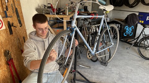 Eine Person trägt einen grauen Hoodie sowie eine blaue Jeans und ist in der Hocke in einer Werkstatt. Die Person schraubt an einem blauen Fahrrad.