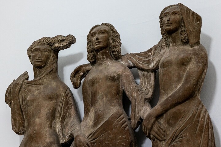 Eine Wandplastik drei weiblicher Figuren, die sich nach vorne blickend an den Händen halten.