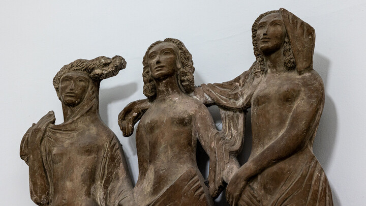 Eine Wandplastik drei weiblicher Figuren, die sich nach vorne blickend an den Händen halten.