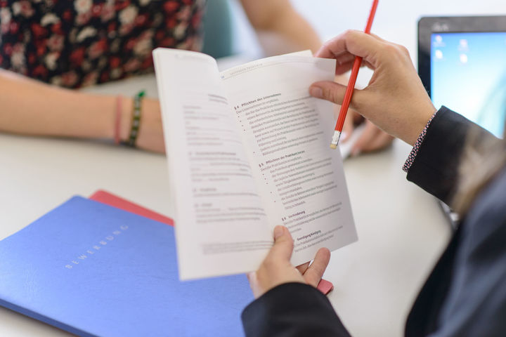 Eine Person hält ein aufgeschlagenes Gesetzbuch in beiden Händen. Darunter liegt eine blaue Mappe. Auf dieser Mappe steht das Wort Bewerbung.