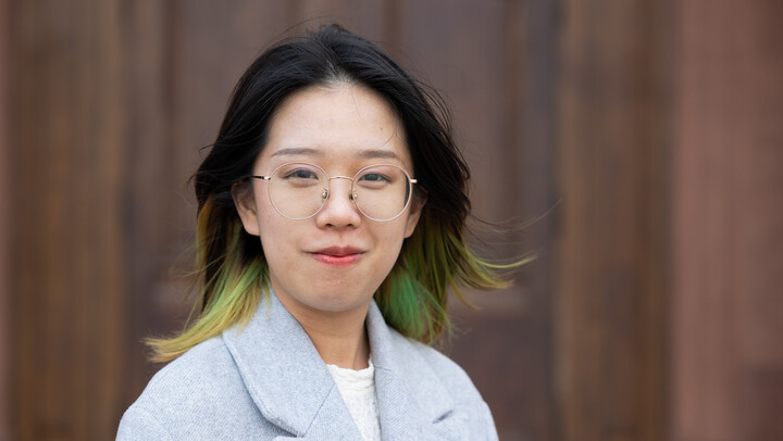 Hongting Jiang. She is wearing a grey jacket over a white shirt and is standing in front of a brown door.