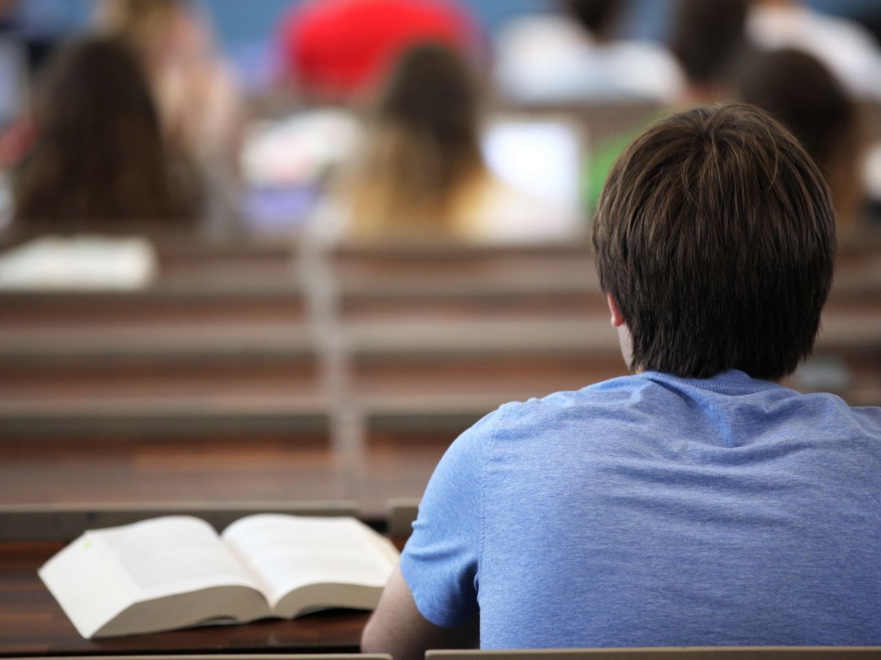 Rückenansicht eines Studierenden mit blauem T-Shirt und braunen Haaren. Er sitzt in einem Hörsaal, vor ihm ein aufgeklapptes Buch.
