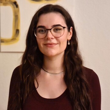 Jule Schabinger. Sie trägt einen schwarzen Pullover und steht vor einer weißen Wand mit einem Gemälde mit gelben Quadraten.
