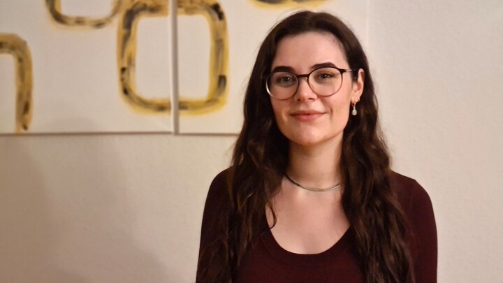 Jule Schabinger. She is wearing a black sweater and is standing in front of a white wall with picture with yellow quadrats.