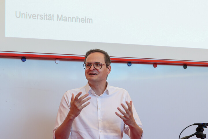 Prof. Dr. Markus Strohmaier, der vor einer Leinwand steht und einen Vortrag hält. Er trägt ein weißes Hemd und hat die Arme gestikulierend gehoben.