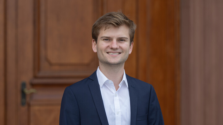 Felix Jungmann. He is wearing a blue suit and white shirt and is standing in front of one of the castle doors.