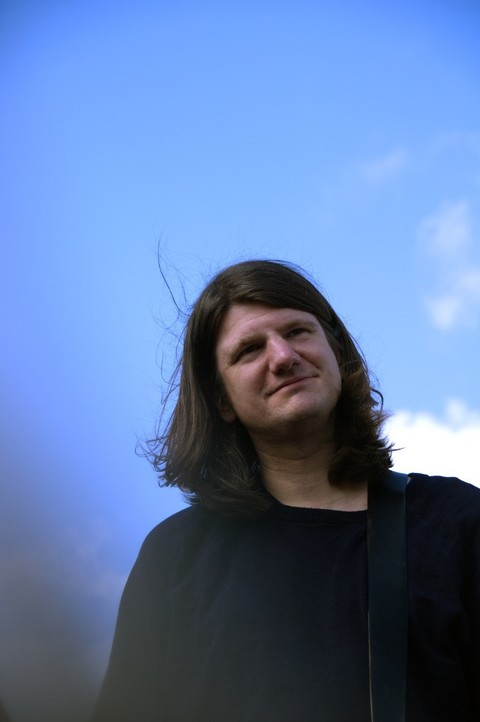 Ein Mann mit langen dunklen Haaren und einem schwarzen Outfit vor blauem Himmel.