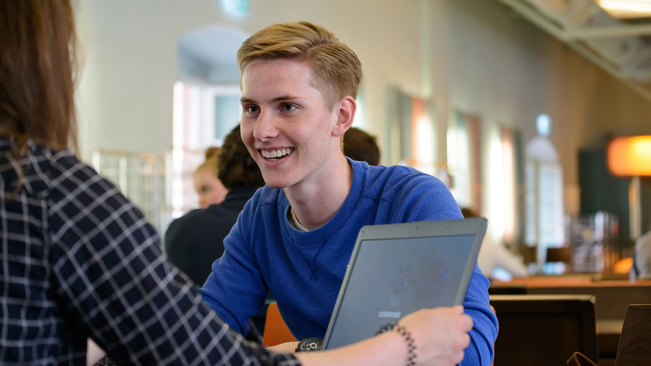 Ein Student und eine Studentin sitzen sich gegenüber an einem Tisch. Der Student trägt kurzes blondes Haar, einen blauen Pullover und lächelt.