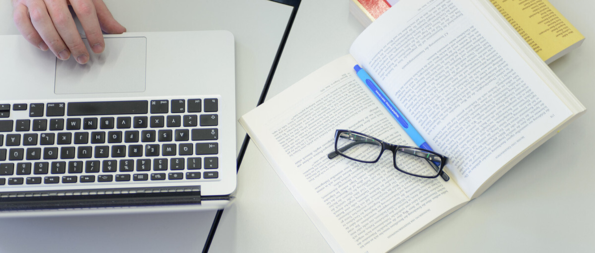 Ein Laptop, ein aufgeschlagenes Buch und eine Brille liegen auf dem Tisch.