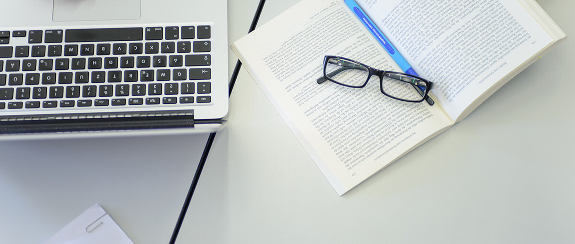 Ein Laptop, ein aufgeschlagenes Buch und eine Brille liegen auf dem Tisch.