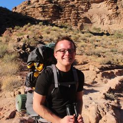 Alexander von Kiesling steht mitten in der Wüste bzw. Dühnen. Er trägt einen großen Wanderrucksack auf seinem Rücken, ein schwarzes T- Shirt, eine graue Wanderhose sowie eine Brille. In den Händen hält er Laufstöcke. Er lächelt.