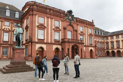 Vier Studierende hören einer Studentin beim Erzählen zu. Sie stehen auf dem Ehrenhof der Universität Mannheim. Im Hintergrund befindet sich das Mannheimer Schloss.