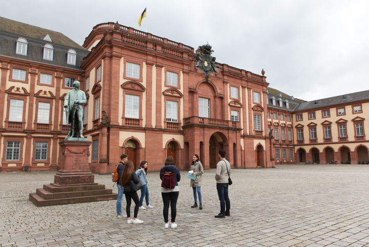 Vier Studierende hören einer Studentin beim Erzählen zu. Sie stehen auf dem Ehrenhof der Universität Mannheim. Im Hintergrund ist das Mannheimer Schloss zu sehen. 