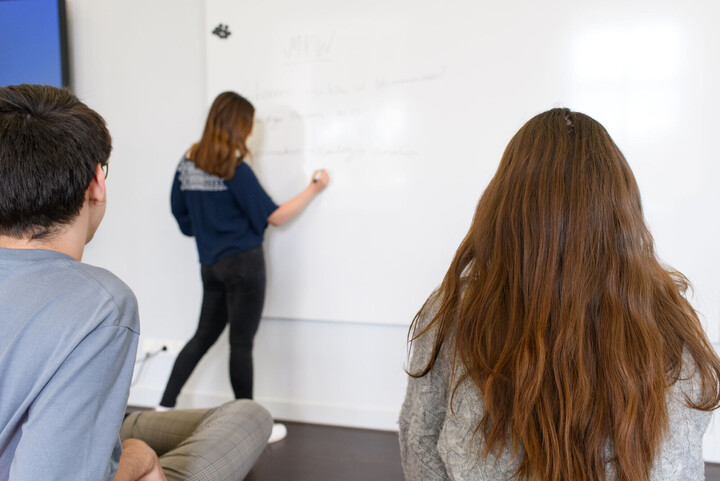 Eine Studentin schreibt auf eine Tafel. Zwei Studierende schauen ihr zu.