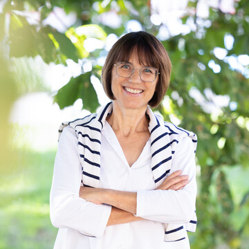  Prof. Dr. Annette Kehnel. Sie steht lächelnd unter einem Baum, dabei trägt sie eine Brille und eine weiße Bluse, ein gestreifter Pullover liegt um ihre Schultern.