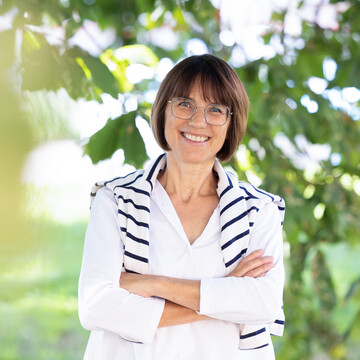  Prof. Dr. Annette Kehnel. Sie steht lächelnd unter einem Baum, dabei trägt sie eine Brille und eine weiße Bluse, ein gestreifter Pullover liegt um ihre Schultern.