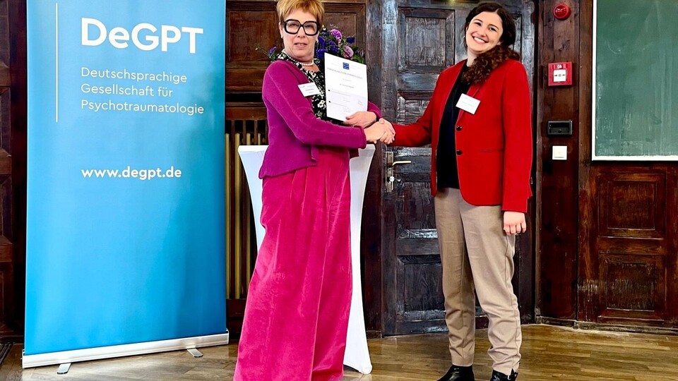 Sarah Danböck (schwarze Haare, rotes Jackett), bekommt eine Urkunde überreicht von einer Frau mit pinkfarbener Hose und Jacke. Beide schauen in die Kamera. Im Hintergrund ist ein Poster der DeGTP zu sehen.