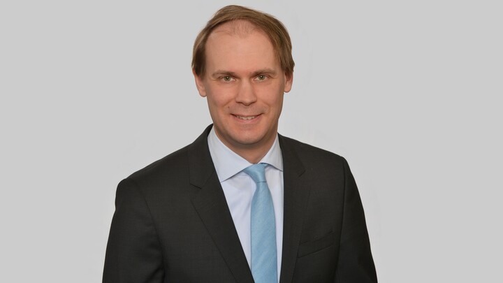 Portrait von Jannis Bischof. Er trägt ein weißes Hemd, eine hellblaue Krawatte und ein schwarzes Sakko.