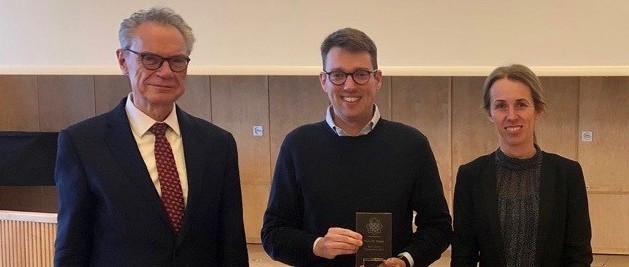 Drei Personen stehen in der Aula, der Mittlere präsentiert einen Preis