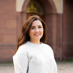 Eine junge Frau steht vor dem Mannheimer Schloss. Sie trägt einen grauen Strickpullover und lange, braune Haare.