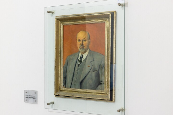 Porträt von Julius Paul Stiegler mit Kinnbart und lichtem Haar, in grauem Anzug mit Weste vor orangefarbenem Hintegrund.