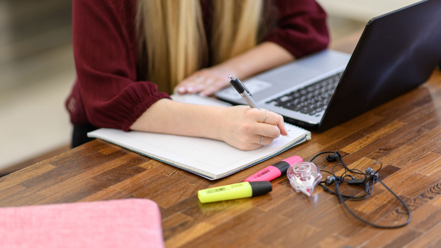 Bildausschnitt einer Person mit langen blonden Haaren und rotem Pullover, die vor einem Laptop sitzt.