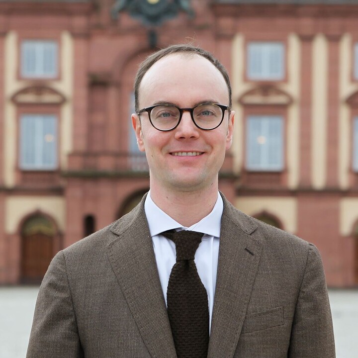 Portrait eines Mannes in Anzug auf dem Ehrenhof. Im Hintergrund das Schloss.
